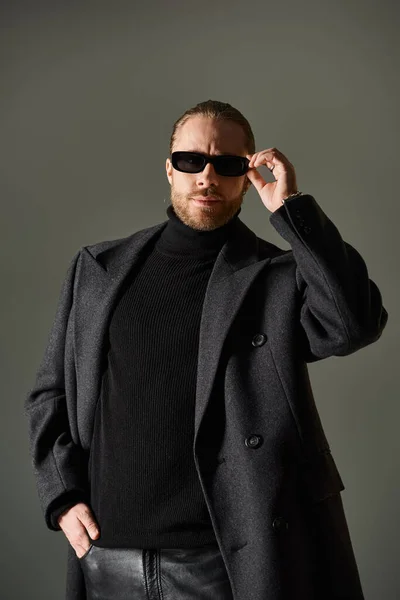 Portrait of fashionable man in trendy sunglasses and coat posing with hand in pocket on grey — Stock Photo