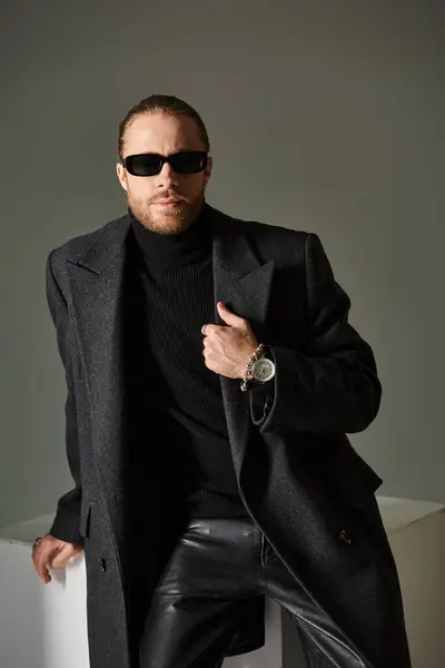 Ritratto di uomo alla moda in occhiali da sole alla moda e cappotto seduto su cubo bianco su sfondo grigio — Foto stock