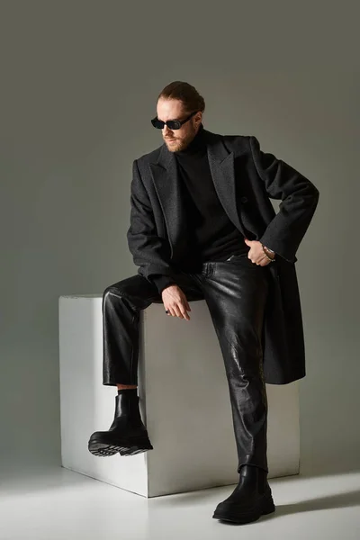 Full length, fashionable model in trendy sunglasses and coat sitting on white cube on grey backdrop — Stock Photo