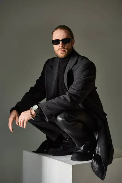 Fashionable male model in trendy sunglasses and black attire sitting on top of on white cube on grey — Stock Photo