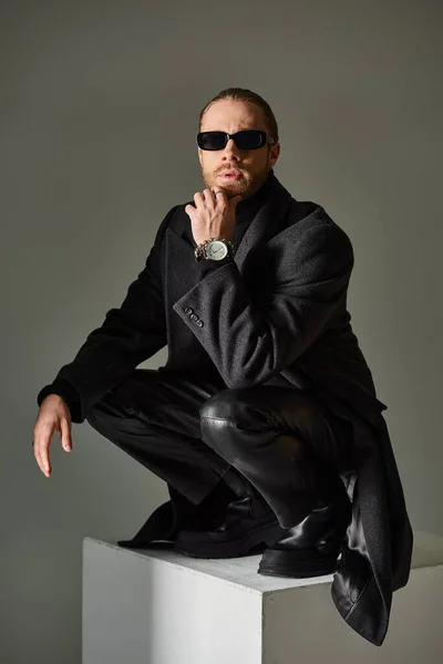 Modelo masculino elegante em óculos de sol na moda e traje preto sentado em cima de no cubo branco no cinza — Fotografia de Stock