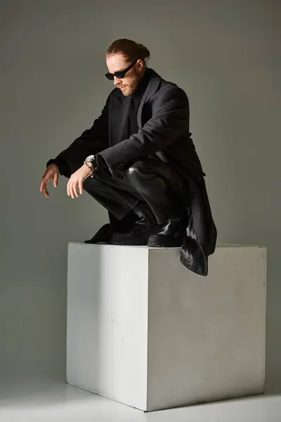 Красивый мужчина модель в модных солнцезащитных очках и черный наряд сидит поверх на белом кубе на сером — стоковое фото