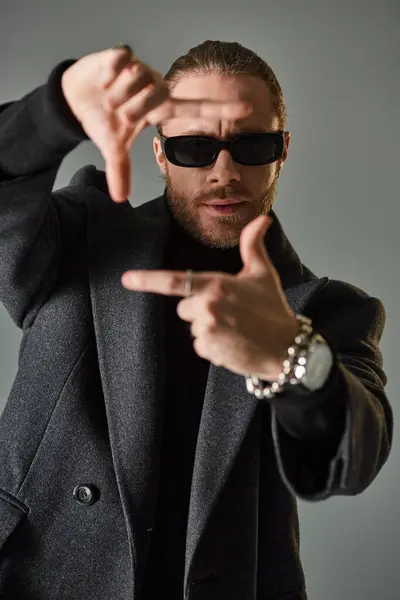 Portrait of handsome male model in trendy sunglasses and black attire showing frame gesture on grey — Stock Photo