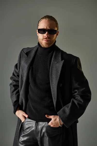Portrait of handsome male model in trendy sunglasses and black attire standing with hands in pockets — Stock Photo