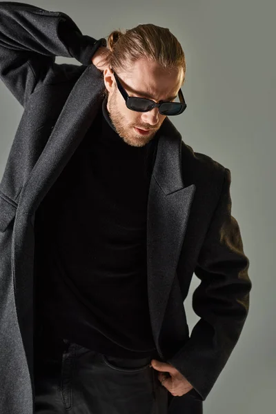 Portrait of handsome male model in trendy sunglasses and black attire standing on grey backdrop — Stock Photo
