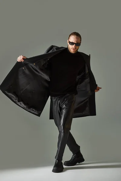 Retrato de hombre barbudo guapo en gafas de sol de moda y traje negro caminando sobre fondo gris - foto de stock
