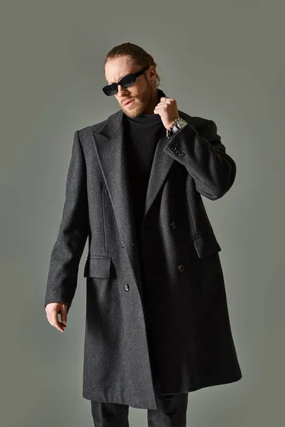Handsome bearded man in trendy sunglasses and black attire adjusting collar of turtleneck — Stock Photo