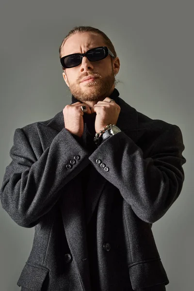 Handsome bearded man in trendy sunglasses and black attire adjusting collar of turtleneck — Stock Photo