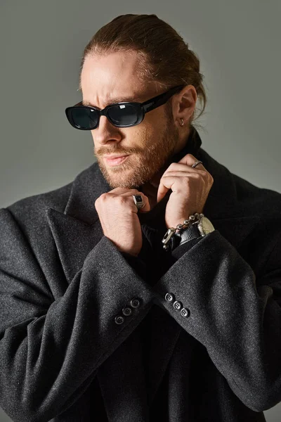 Homem barbudo de boa aparência em óculos de sol na moda e traje preto ajustando colar de gola alta — Fotografia de Stock