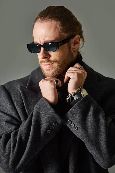 Good looking bearded man in trendy sunglasses and black attire adjusting collar of turtleneck — Stock Photo