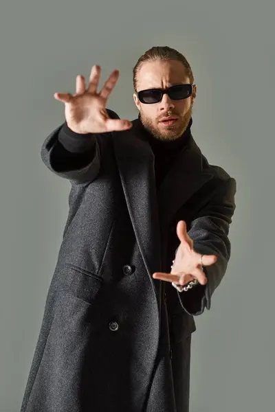 Good looking bearded man in trendy sunglasses and black attire posing and gesturing on grey — Stock Photo
