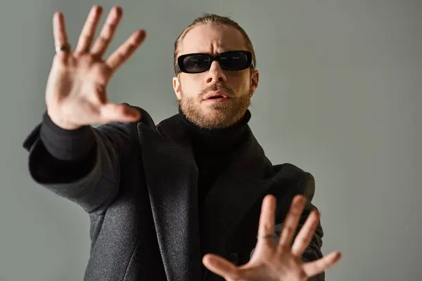 Good looking bearded model in trendy sunglasses and black attire posing and gesturing on grey — Stock Photo