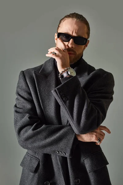 Stylish and bearded man in trendy dark sunglasses and black attire posing on grey background — Stock Photo