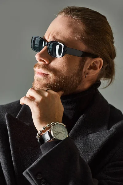 Pensive and bearded man in trendy dark sunglasses and black attire posing on grey background — Stock Photo