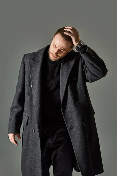 Bello e barbuto uomo in abito nero alla moda e cappotto di lana in posa su sfondo grigio — Foto stock