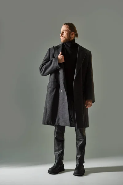 Good looking and bearded man in trendy black attire and woolen coat looking away on grey background — Stock Photo