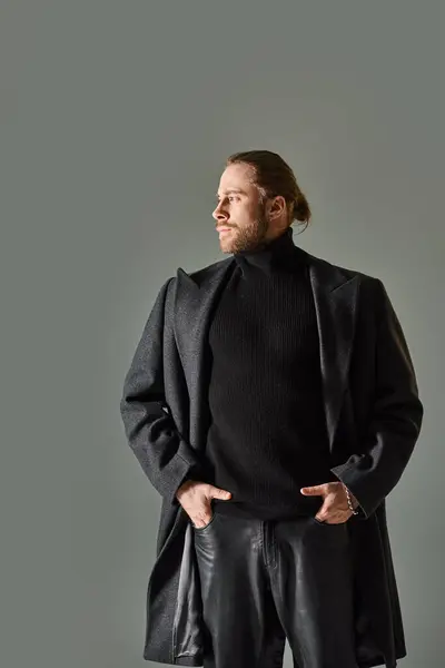 Красивый и бородатый мужчина в модном черном одеянии и шерстяном пальто, смотрящий в сторону на сером фоне — стоковое фото