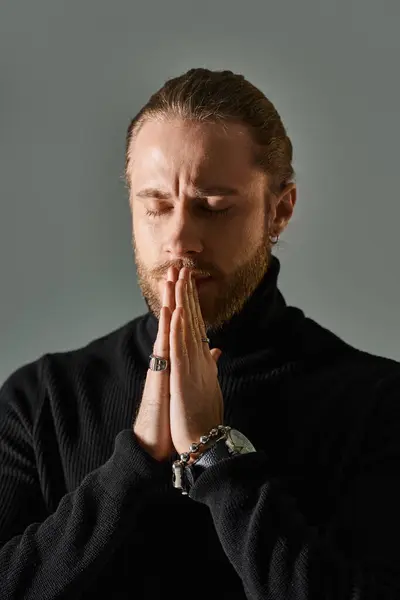 Portrait of handsome man in black turtleneck sweater standing with praying hands on grey background — Stock Photo