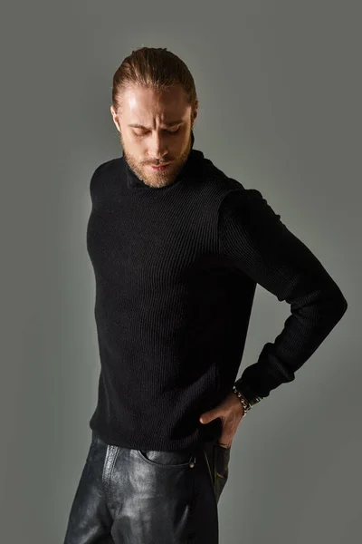 Handsome man in turtleneck sweater posing with hand in pocket of leather pants posing on grey — Stock Photo