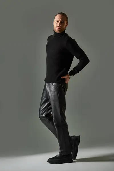 Tutta la lunghezza dell'uomo in maglione dolcevita in posa con mano in tasca di pantaloni in pelle su grigio — Foto stock