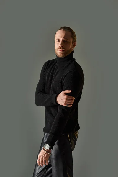 Uomo bello barbuto in maglione collo alto nero e pantaloni di pelle in posa su sfondo grigio — Foto stock