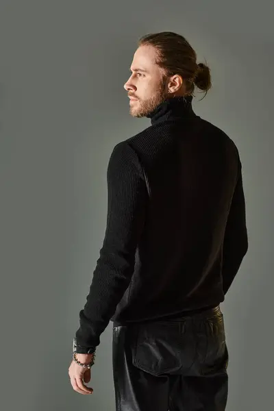 Bearded handsome man in black turtleneck sweater and leather pants standing on grey background — Stock Photo