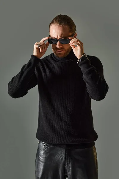 Bearded man in trendy sunglasses, turtleneck sweater and black leather pants posing on grey — Stock Photo