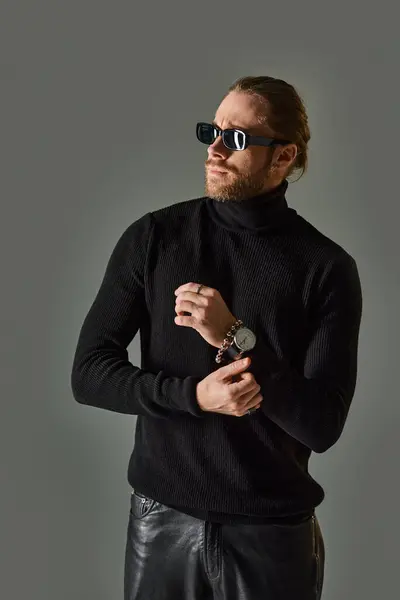 Uomo barbuto in occhiali da sole alla moda, maglione dolcevita e pantaloni in pelle nera in posa su grigio — Foto stock
