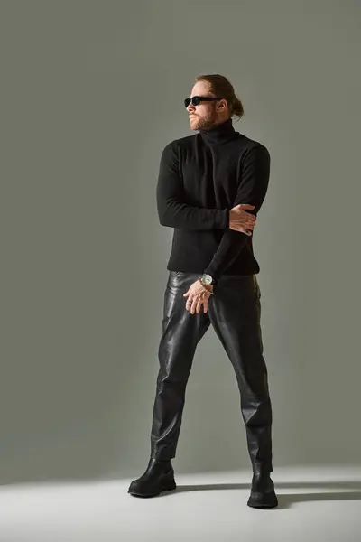 Homme barbu en lunettes de soleil tendance, pull col roulé et pantalon en cuir posé sur gris — Photo de stock