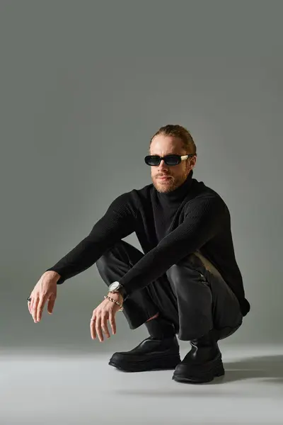Bearded man in trendy sunglasses, turtleneck sweater and leather pants sitting on haunches on grey — Stock Photo