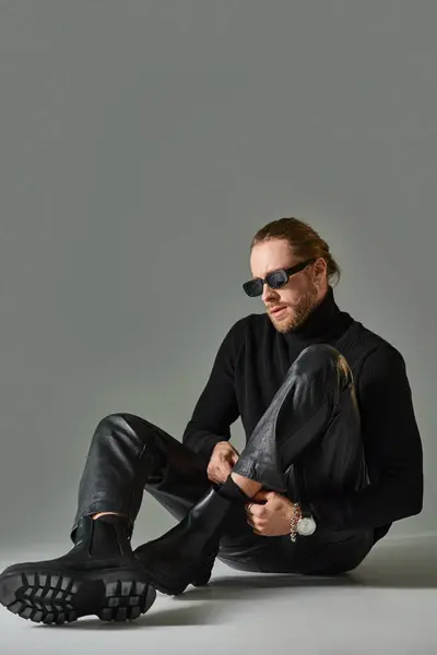 Bearded man in trendy sunglasses, turtleneck sweater and leather pants sitting on grey backdrop — Stock Photo