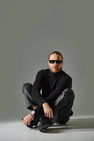 Homme barbu en lunettes de soleil, pull col roulé et pantalon en cuir assis avec jambes croisées sur gris — Photo de stock