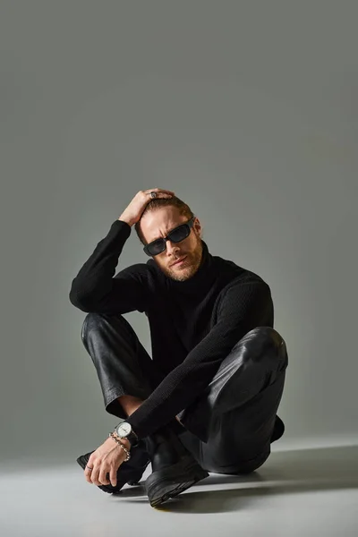 Bearded man in sunglasses, turtleneck sweater and leather pants sitting with crossed legs on grey — Stock Photo
