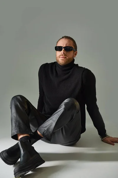 Hombre guapo en gafas de sol, jersey de cuello alto y pantalones de cuero sentado con las piernas cruzadas en gris - foto de stock