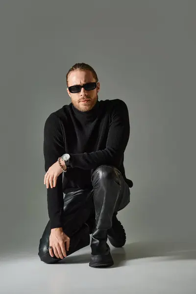 Bearded man in trendy sunglasses, turtleneck sweater and leather pants sitting on grey background — Stock Photo
