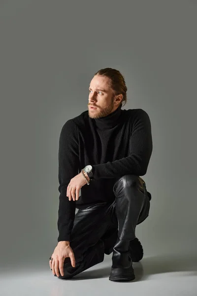 Longitud completa del hombre barbudo en jersey de cuello alto y pantalones de cuero negro sentado sobre fondo gris - foto de stock