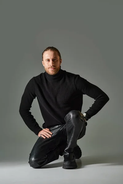 Full length of bearded man in turtleneck sweater and black leather pants sitting on grey background — Stock Photo