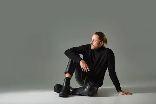 Полная длина бородатый мужчина в водолазке свитер и черные кожаные брюки сидя на сером фоне — стоковое фото
