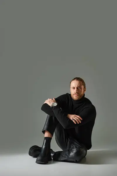 Full length of good looking man in turtleneck sweater and leather pants sitting on grey background — Stock Photo