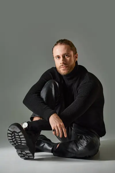 Full length of bearded man in turtleneck sweater and black leather pants sitting on grey backdrop — Stock Photo