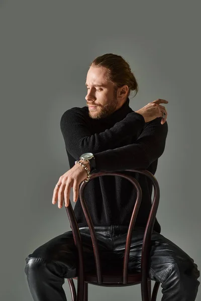 Bearded man in turtleneck and black leather pants sitting on wooden chair on grey background — Stock Photo