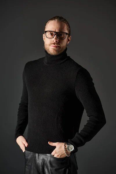 Bearded man in turtleneck sweater and eyeglasses standing and looking at camera on grey background — Stock Photo