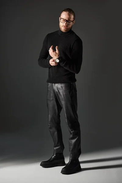 Bearded man in turtleneck sweater and eyeglasses standing in leather pants on grey background — Stock Photo