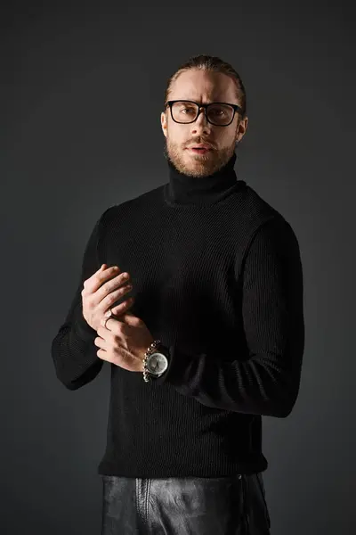 Bearded man in turtleneck sweater and eyeglasses standing in leather pants on grey background — Stock Photo