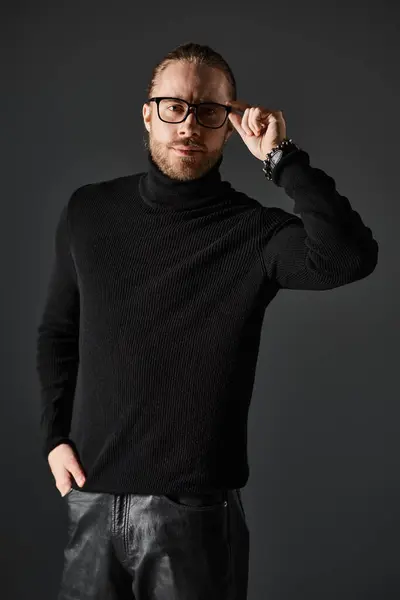 Handsome man in turtleneck sweater and eyeglasses standing in leather pants on grey background — Stock Photo