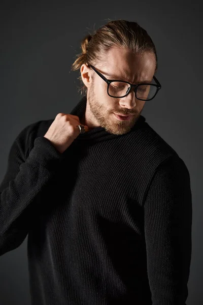 Handsome man in eyeglasses pulling high collar on turtleneck sweater while posing on grey background — Stock Photo