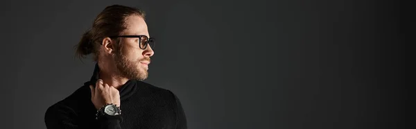 Handsome man in eyeglasses pulling high collar on turtleneck sweater while posing on grey, banner — Stock Photo
