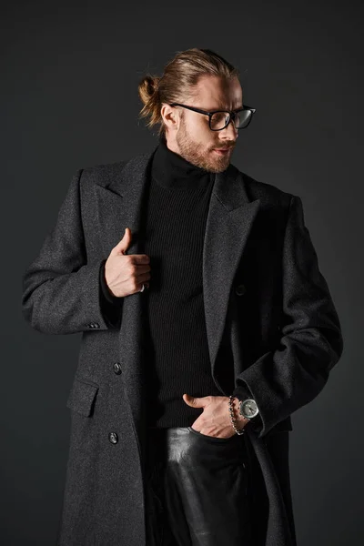 Hombre barbudo con gafas y elegante abrigo de lana posando con la mano en el bolsillo sobre fondo gris - foto de stock