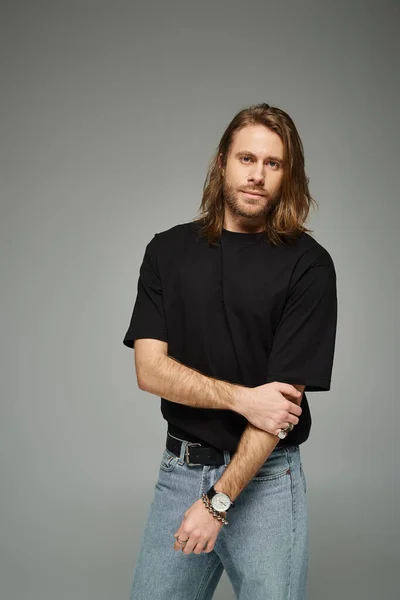 Bello uomo barbuto con i capelli lunghi in piedi in casual t-shirt e jeans su sfondo grigio — Foto stock