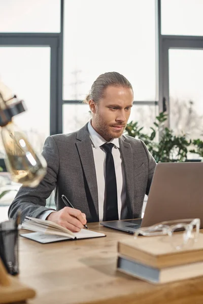 Homem de negócios concentrado escrevendo em notebook e olhando para laptop no local de trabalho no escritório moderno — Fotografia de Stock